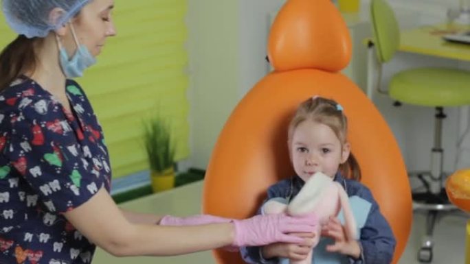 牙医女孩安抚坐在牙科椅上的小女孩给孩子一个柔软的玩具。