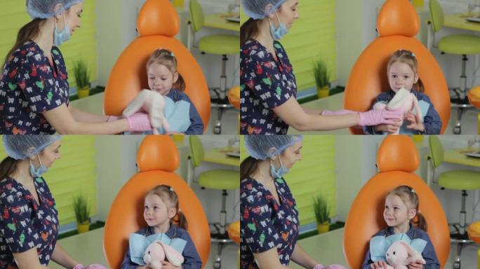 牙医女孩安抚坐在牙科椅上的小女孩给孩子一个柔软的玩具。