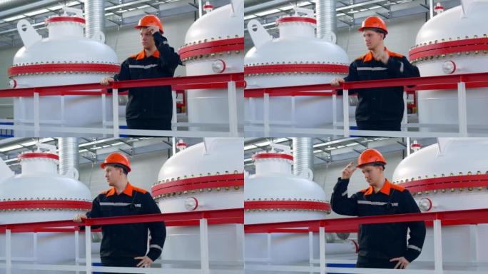 一名戴着橙色头盔的年轻工厂工人站在煤气罐附近的煤气液化店里，调整他的工作头盔。石油天然气工业和石油运