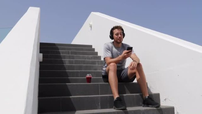 男子听音乐手机app戴着耳机坐在城市楼梯上。健康的城市生活方式的人在户外慢跑时使用智能手机，早晨的红