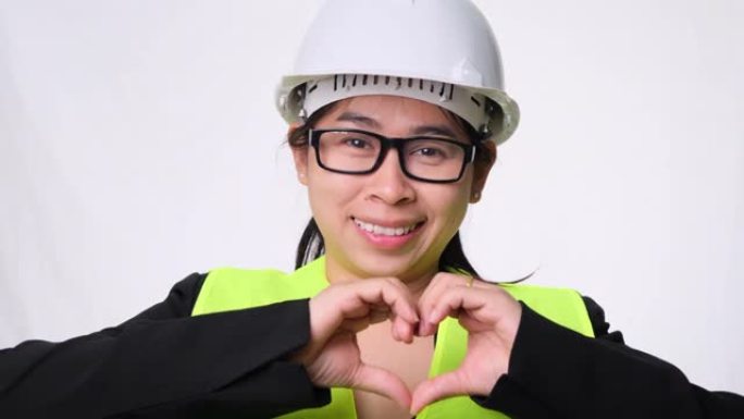 在工作室里，戴着头盔的女工程师带着头盔微笑，双手在白色背景上展示心形。