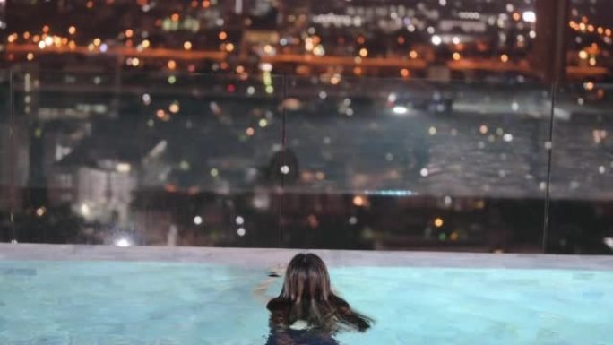 快乐的广角年轻女子在室外游泳池，晚上可以看到城市的景色