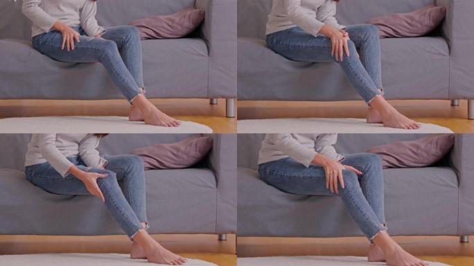 一个女人坐在灰色的沙发上，试图在大腿和关节上做自我按摩。她挤压肌肉以减轻痛苦。按摩可以帮助她从疼痛中