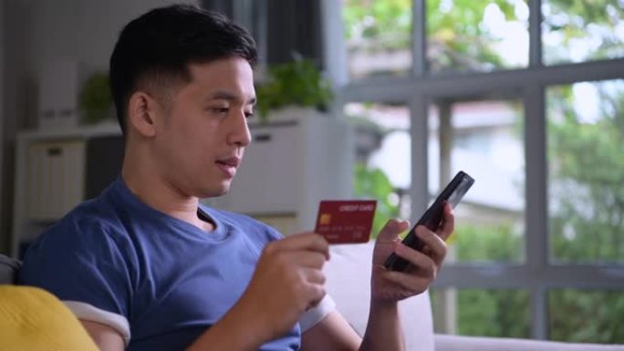 亚洲男子舒适地坐在家里享受和浏览网上购物，使用信用卡和智能手机上的在线支付和银行应用程序轻松便捷地支
