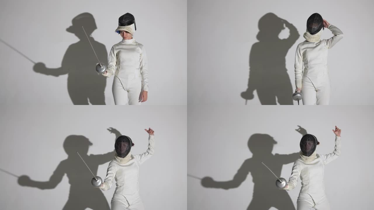 一名年轻女子击剑手的肖像用剑杆敬礼，戴上头盔，进入战斗位置。运动员在带有阴影的白色背景上的黑暗工作室