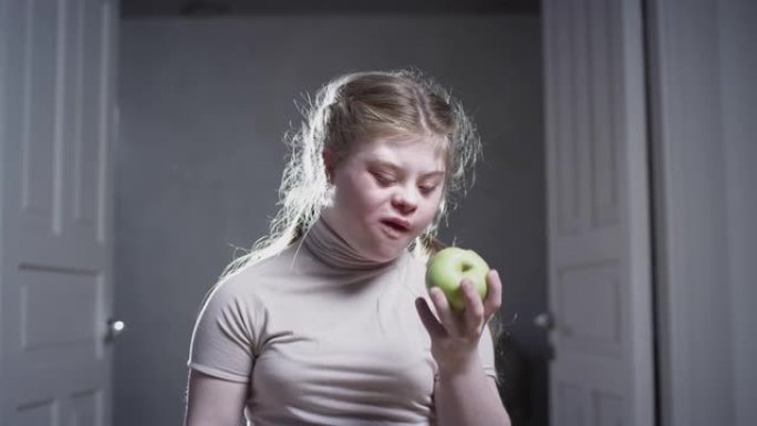 一个患有唐氏综合症的女孩正在咬一个青苹果。健康食品。残疾人在家。残疾生活