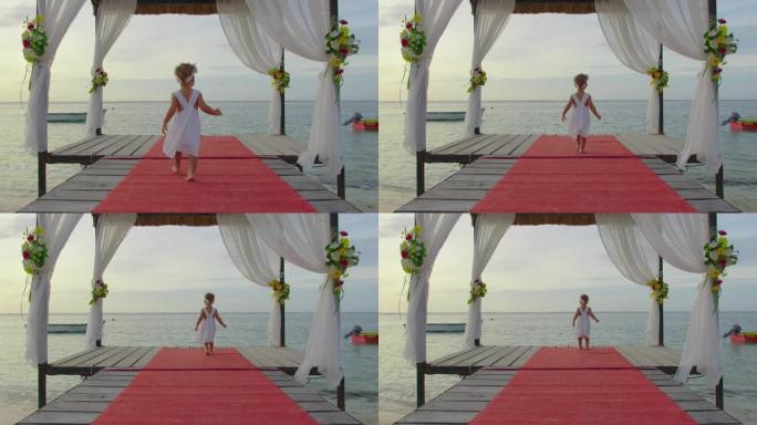 日落时分，印度洋沿岸，一个穿着白色连衣裙的两岁小女孩沿着鲜花装饰的码头散步。婚礼视频概念