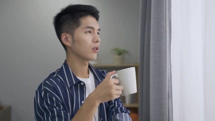 穿着休闲装的快乐亚洲男人在一个宁静的早晨，在明亮的家庭室内，从杯子里喝一杯热饮料时，轻柔地叹了口气。