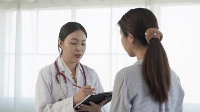 4K，一名白人亚裔女医生站在医院检查室，对一名身穿长袖衬衫的长发亚裔患者做笔记。概念健康与医学