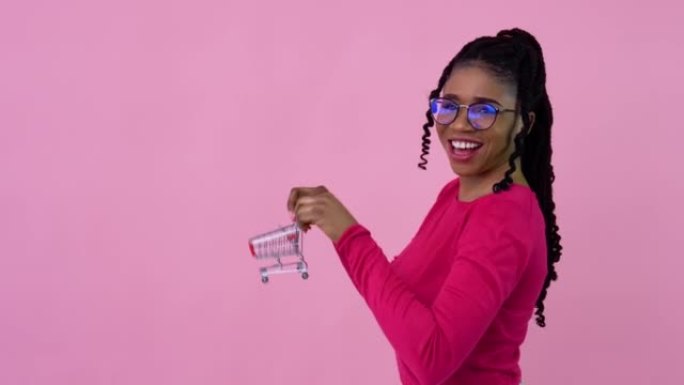 穿着粉色衣服的可爱开朗的年轻非洲裔美国女孩拿着玩具迷你购物车。少女家庭主妇初学者站在坚实的粉红色背景