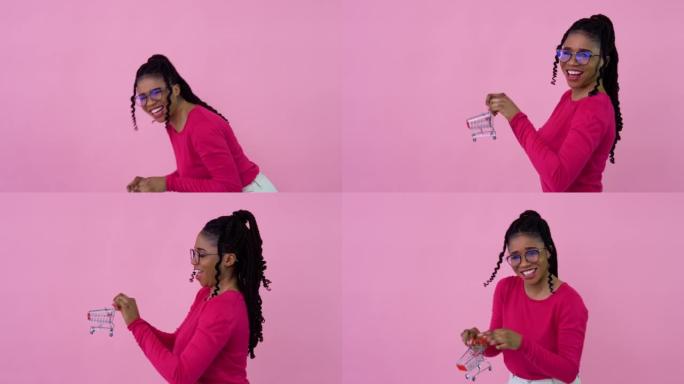 穿着粉色衣服的可爱开朗的年轻非洲裔美国女孩拿着玩具迷你购物车。少女家庭主妇初学者站在坚实的粉红色背景