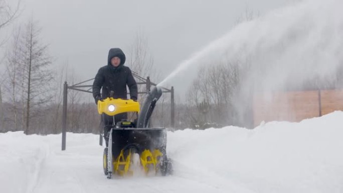 一名男子在院子里用吹雪机在慢动作降雪时清理积雪。前视图。慢动作