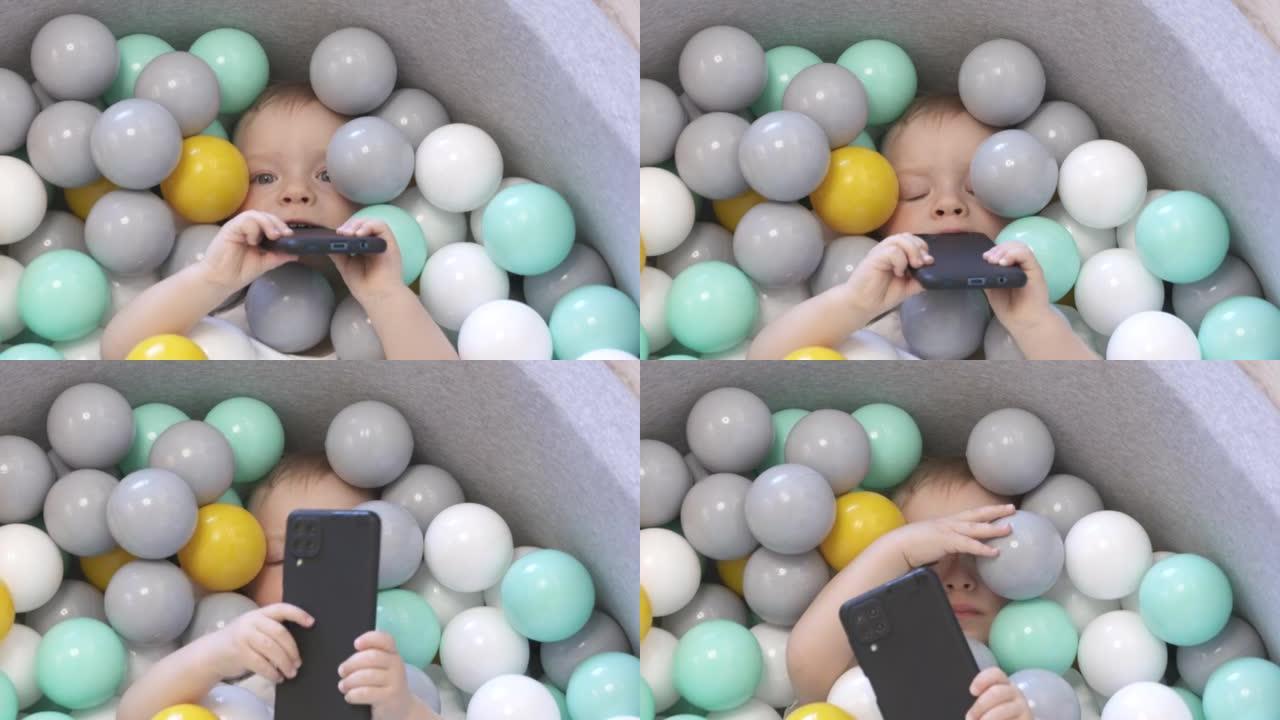 孩子在家里的球坑里玩智能手机。快乐的孩子躺在软球坑里，有五颜六色的球给幼儿，高加索男婴在家里玩得开心