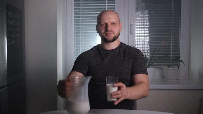 一个英俊的秃头运动男子将玻璃水瓶中的牛奶倒入玻璃杯中，并在黑暗中在厨房里喝酒
