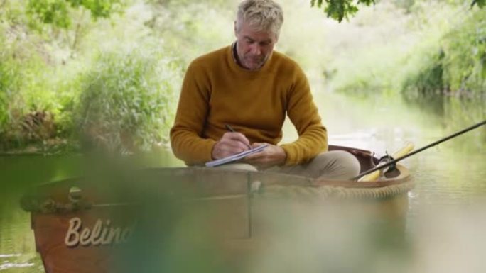 成熟的男人在河上的木船上钓鱼时在笔记本上写字