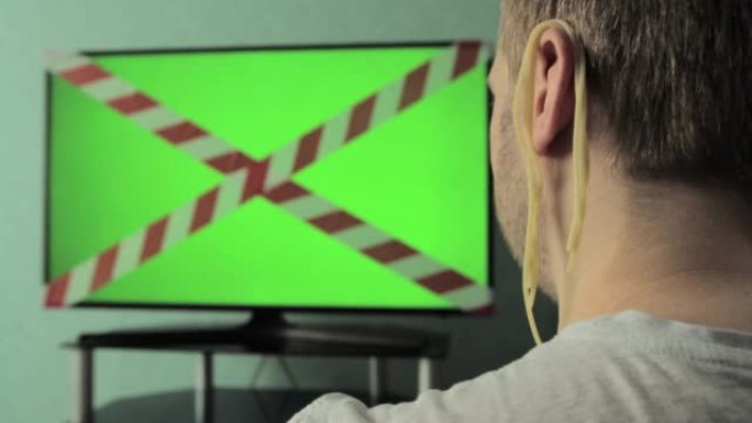 一个耳朵上有面条的男人正在看电视，上面有一个色键绿色屏幕和一个禁止磁带。电视和互联网上的假货概念，制