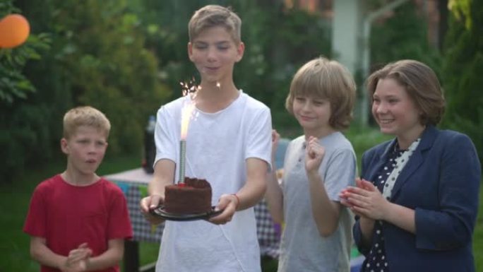 快乐的十几岁的男孩，在生日蛋糕上燃烧着蜡烛，站在后院，朋友们在慢动作中鼓掌微笑。自信的高加索少年在户