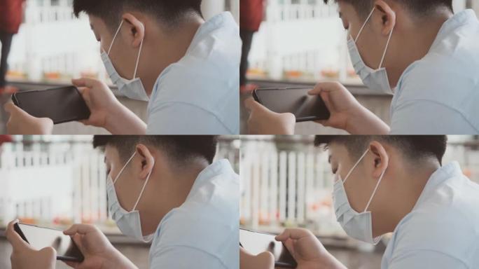 亚洲男孩戴着口罩，坐在百货商店玩手机游戏，新常态和社交疏远的生活方式概念。