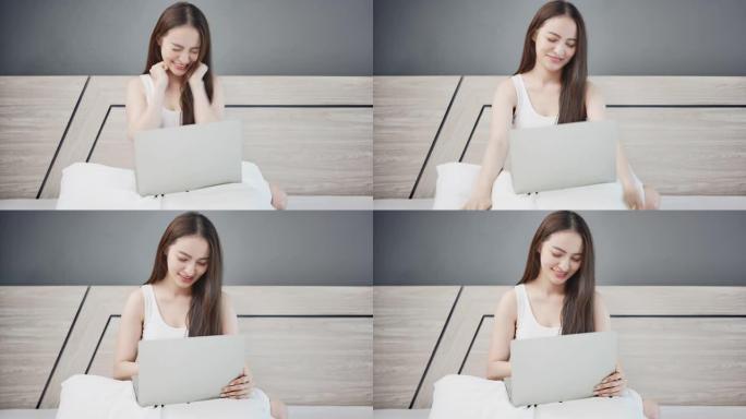 年轻的亚洲妇女坐在和使用笔记本电脑在线学习，并在家里的卧室床上得到一个好消息。在家工作的概念