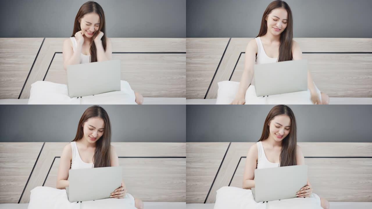 年轻的亚洲妇女坐在和使用笔记本电脑在线学习，并在家里的卧室床上得到一个好消息。在家工作的概念