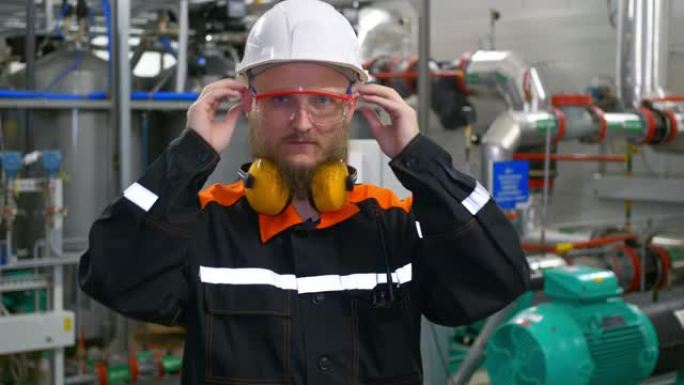 一位戴着白色头盔的大胡子工业工程师站在生产车间的工作场所，脱下防护耳机和护目镜。重工业工人看着镜头