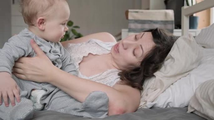 一位穿着睡衣的白人黑发母亲和她的小新生儿一起醒来，在床上放松。带着婴儿睡在家里的年轻女子。同床概念。