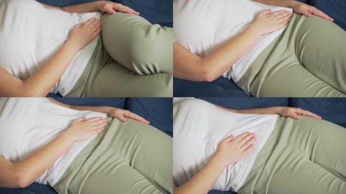 穿着白色t恤的女人躺在蓝色沙发上，用手托着肚子。用手抚摸胃，这样就不会受伤。月经，怀孕，腹泻，暴食。