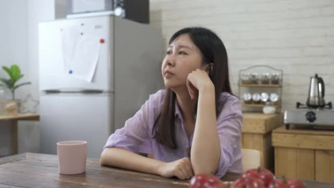 沮丧的亚洲妇女在家里的餐桌旁望着远处时，发出沉重的叹息，并在沉思中摇了摇头。