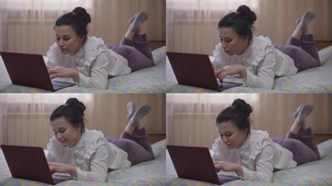 宽镜头放松积极的年轻女子躺在床上在社交媒体上在线传递信息。快乐无忧无虑的千禧一代高加索女士在家里享受