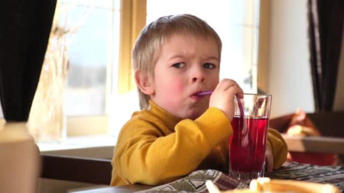 金发男孩婴儿在咖啡馆喝柠檬水。3岁的孩子通过吸管从玻璃杯中喝红汁。在餐馆里蹒跚学步的男孩。特写自然情