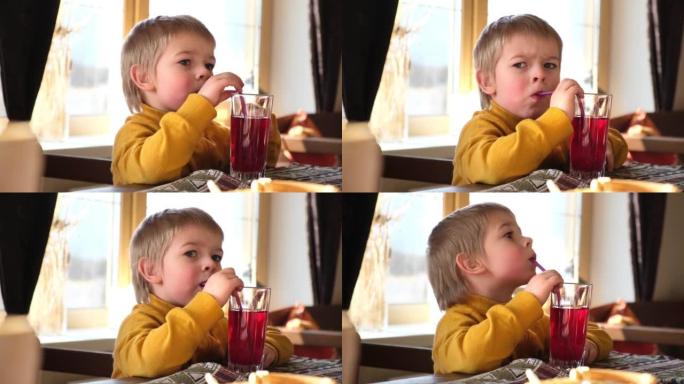 金发男孩婴儿在咖啡馆喝柠檬水。3岁的孩子通过吸管从玻璃杯中喝红汁。在餐馆里蹒跚学步的男孩。特写自然情