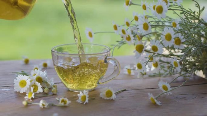 洋甘菊茶从透明茶壶倒入玻璃杯中。动作在夏季乡村露台上进行，并播放阳光。附近是药用洋甘菊的花。