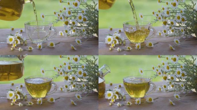 洋甘菊茶从透明茶壶倒入玻璃杯中。动作在夏季乡村露台上进行，并播放阳光。附近是药用洋甘菊的花。