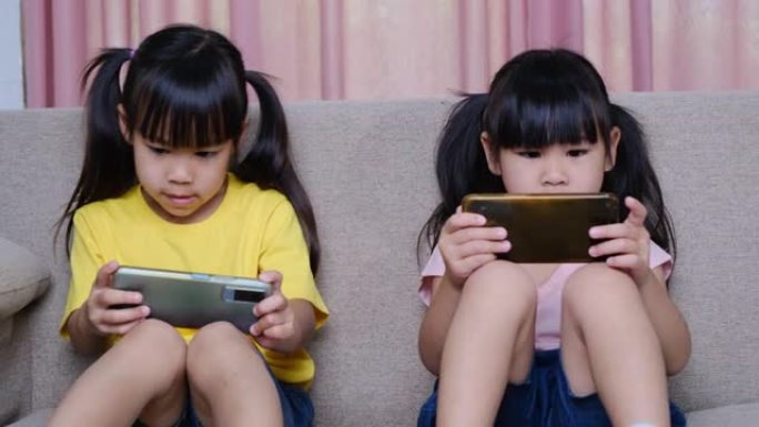 两姐妹坐在家里的沙发上，用智能手机玩网络游戏。现代通信和小工具成瘾概念。两个带着小玩意的孩子。