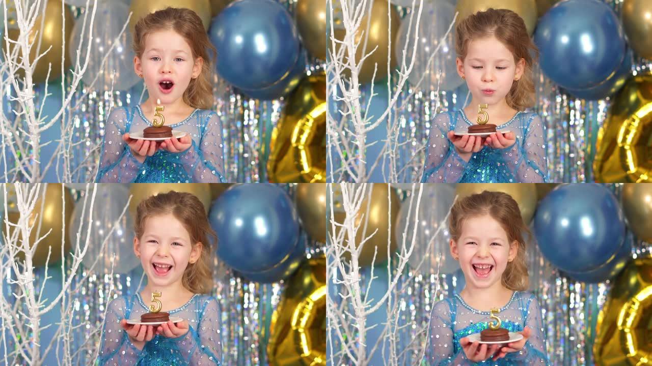 快乐5年。女孩在蓝色背景的纸杯蛋糕中吹出蜡烛。生日那天开朗的孩子。穿着喜庆的蓝色闪亮连衣裙的高加索女