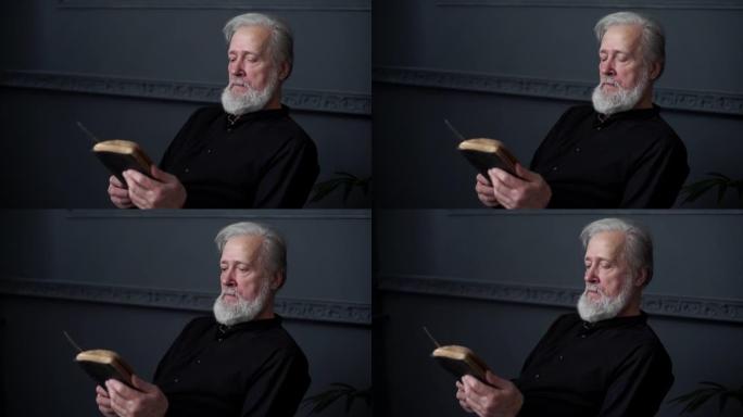 大胡子老人读旧纸质书坐在黑暗的客厅的椅子上，慢动作。