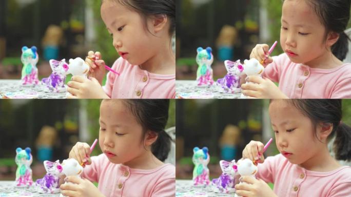 可爱的亚洲女孩小孩蹒跚学步，在后院的房子里专注于灰泥娃娃绘画。通过做艺术品来发展儿童小肌肉。
