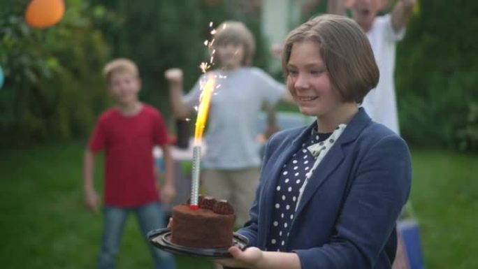 兴奋的高加索少女欣赏生日蜡烛在蛋糕中燃烧，一群模糊的朋友在背景中欢欣鼓舞。快乐少年在后院户外庆祝节日
