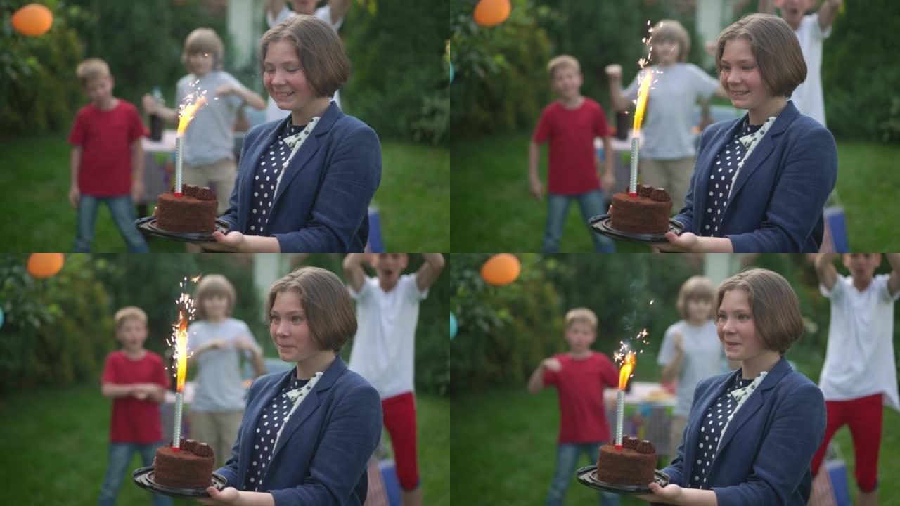 兴奋的高加索少女欣赏生日蜡烛在蛋糕中燃烧，一群模糊的朋友在背景中欢欣鼓舞。快乐少年在后院户外庆祝节日