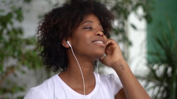 开朗的非洲年轻女子的特写脸戴上耳机，开始在绿色植物的灯光室里听最喜欢的音乐。