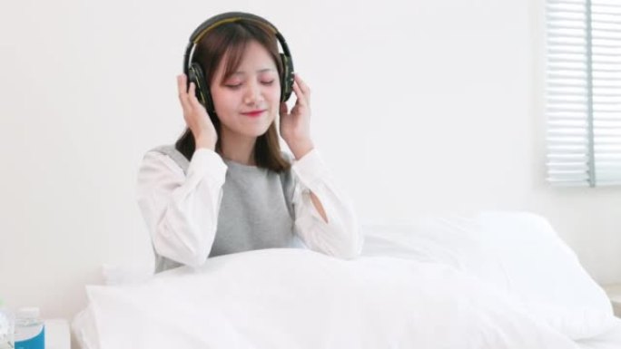 年轻的亚洲女子在白色卧室的床上戴着无线耳机跳舞和听音乐。感到快乐。