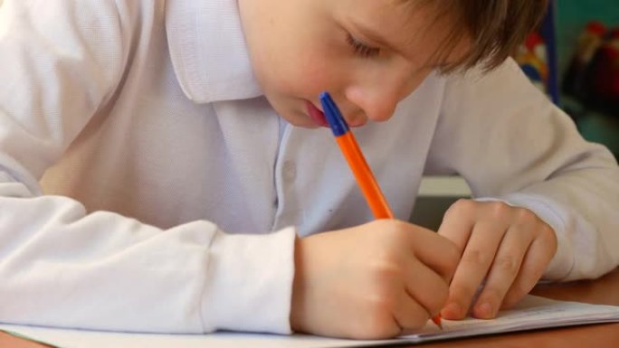 7岁的白人男孩坐在桌子旁用笔在笔记本上写字。这个男孩学会了写大写字母。学生做作业，在笔记本上写字。家