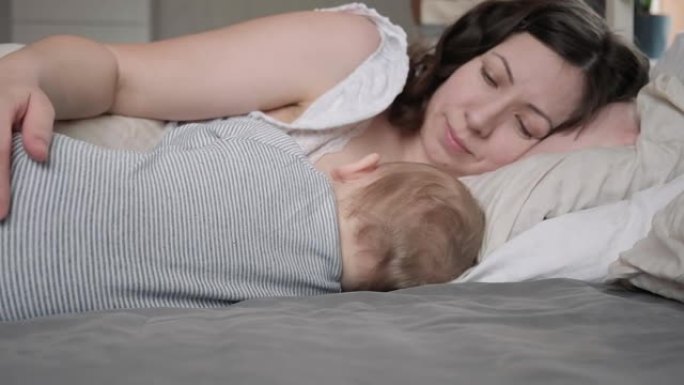 一位穿着睡衣的白人黑发母亲在床上放松时母乳喂养她的小新生儿。带着婴儿睡在家里的年轻女子。同床概念。婴