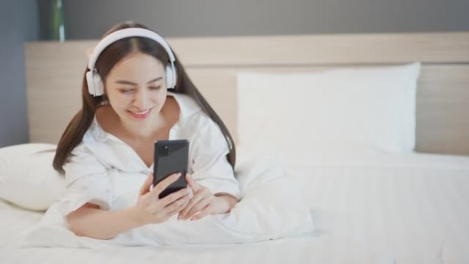 美丽的亚洲女人用智能手机听耳机音乐，和男朋友聊天，在卧室舒适休息的床上微笑快乐酒店人的生活方式