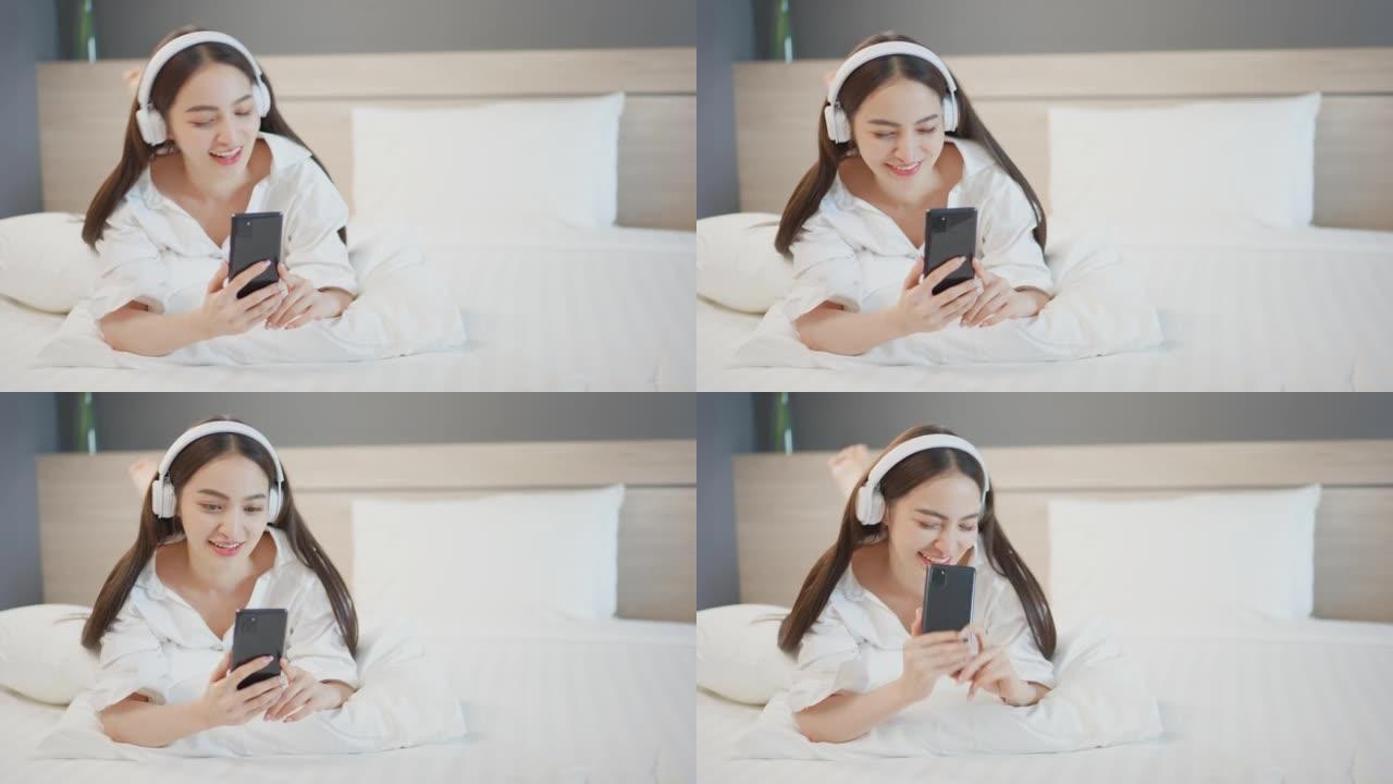 美丽的亚洲女人用智能手机听耳机音乐，和男朋友聊天，在卧室舒适休息的床上微笑快乐酒店人的生活方式