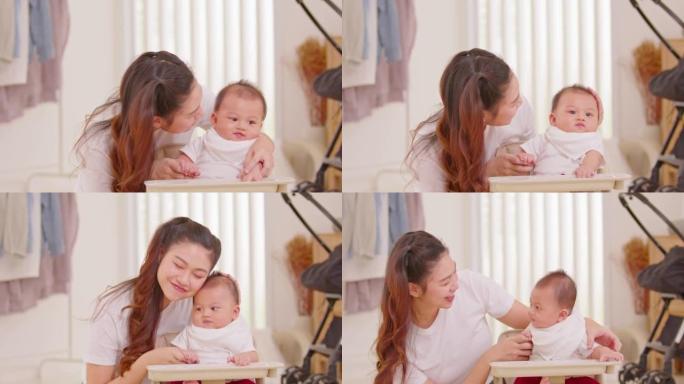 新的亚洲妈妈在椅子上扮演可爱的新生婴儿，在家里微笑和幸福。快乐的母亲与婴儿交谈，用爱亲吻婴儿脸颊。婴