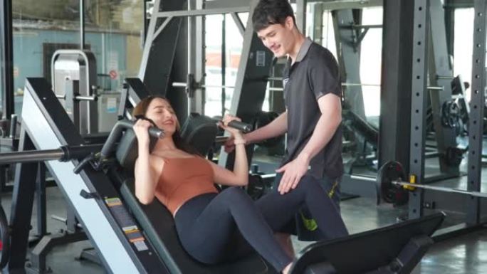 亚洲健身教练男性支持，教和激励年轻美丽的初学者运动员通过举重锻炼机器锻炼。积极的健美女孩运动，在体育