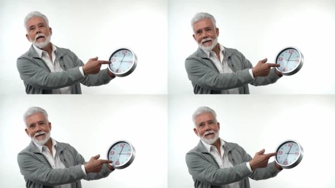 一位白人时尚老人的肖像指向时钟概念时间就是金钱。在白色背景的工作室里。趋势老化的概念。