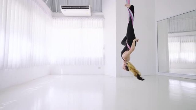 穿着黑色运动服的女运动员，练习飞行瑜伽体式，反重力瑜伽练习，用一条腿撑住倒立伸展，用吊床把另一条腿向