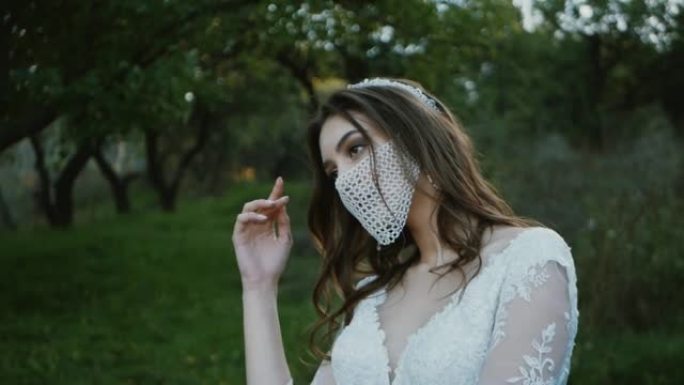 戴着面具摆姿势的新娘肖像。公园里穿着婚纱的漂亮年轻新娘。她的脸上有一个带鹅卵石的时尚白色面具